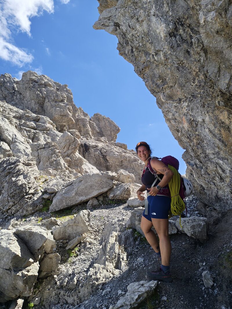 Emma smiling on a rocky mountain trail, carrying a climbing rope and helmet